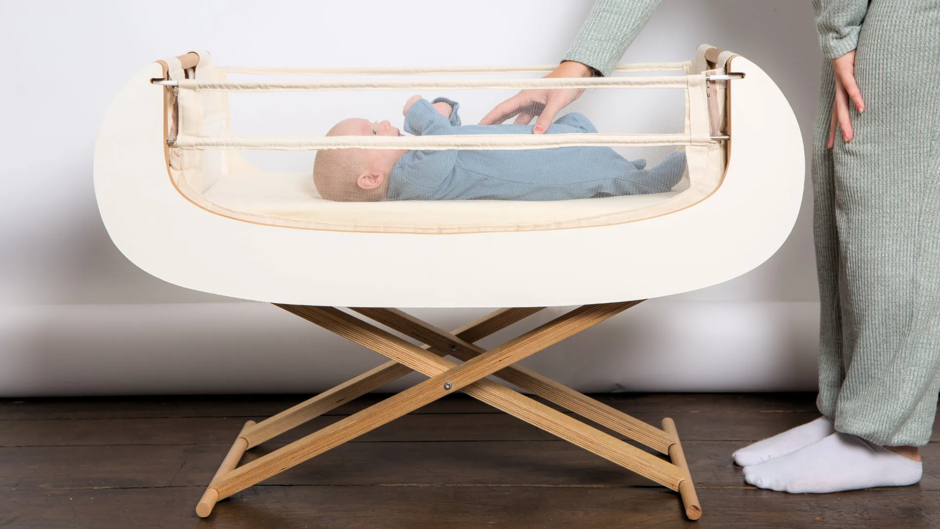 A parent reaching into the Alora bedside crib to settle a baby lying on the mattress, the crib standing on its folding wooden scissor frame.