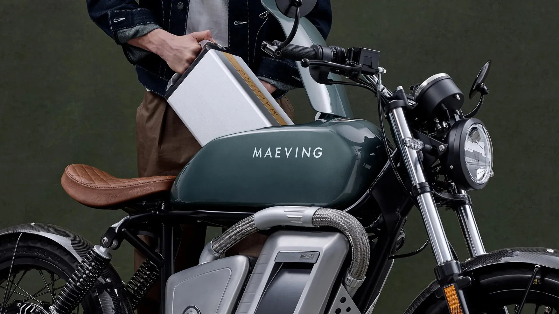 A rider in a denim jacket lifting the removable silver battery pack out of the frame of a sage green Maeving motorcycle.